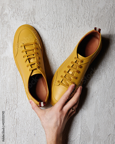 Male hand holding yellow leather sneakers on gray background photo