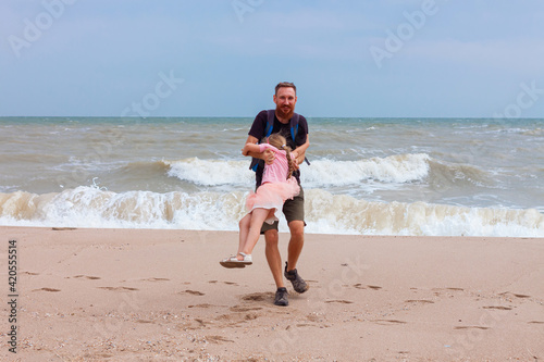 Father holds his cute daughter in arms, circles around himself, play and have fun together on sea beach. Little flying girl smiling hugging with dad outdoor. Loving child and daddy.Father's Day family