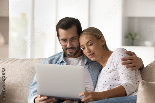 Happy young Caucasian couple relax on sofa at home look at laptop screen watch video online together. Millennial man and woman rest on couch use computer browsing internet or shopping online.