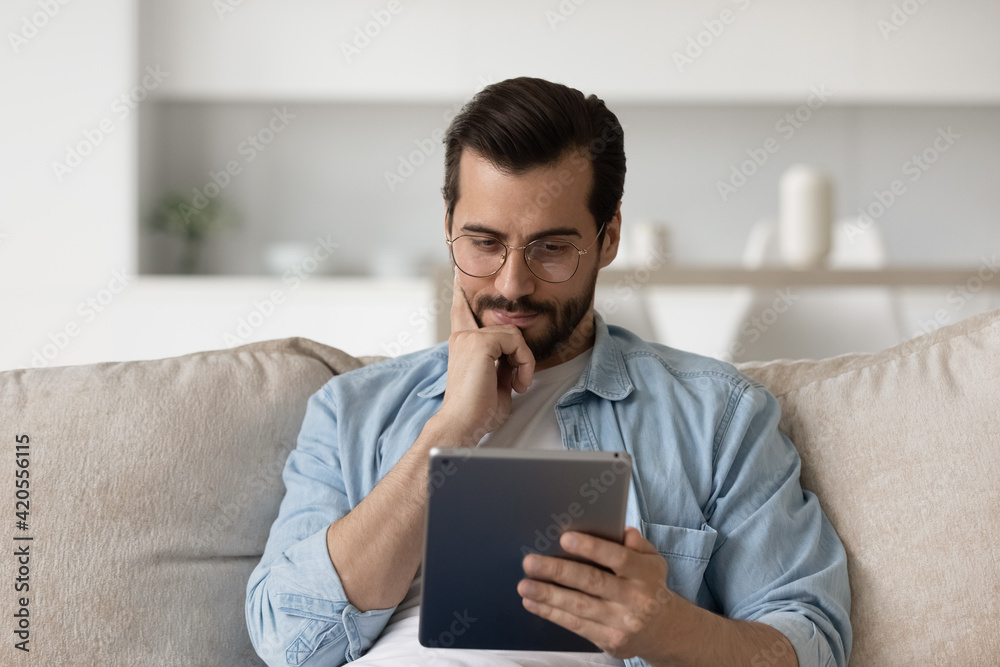 Pensive millennial Caucasian man relax on sofa at home use tablet thinking pondering. Unhappy thoughtful young male look at pad screen consider problem solution on gadget. Technology concept. Pensive millennial Caucasian man relax on sofa at home use tablet thinking pondering. Unhappy thoughtful young male look at pad screen consider problem solution on gadget. Technology concept.