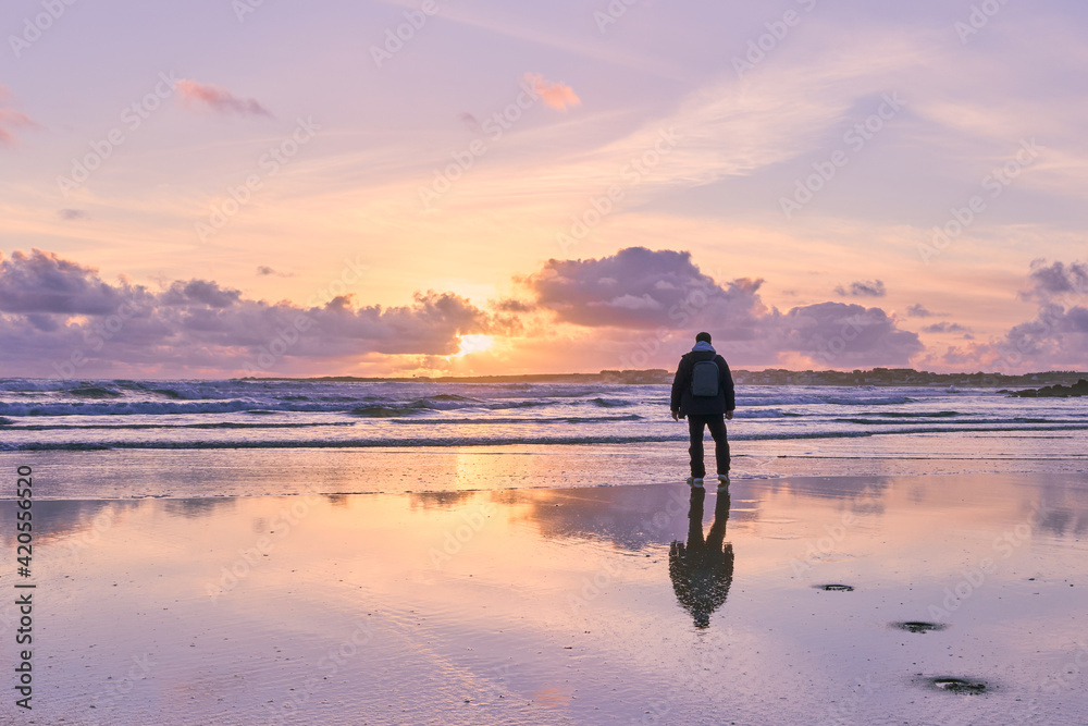 hombre vestido de espaldas en una playa cerca del mar mirando la puesta de sol con colores pastel en invierno. precioso paisaje con el horizonte atravesado por una persona que está mirando el atardece