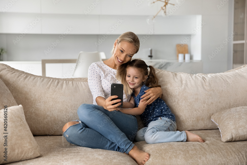 Smiling young Caucasian mother and little 7s daughter relax on sofa at home talk on video call on smartphone. Happy mom and small girl child use cellphone make self-portrait picture together.