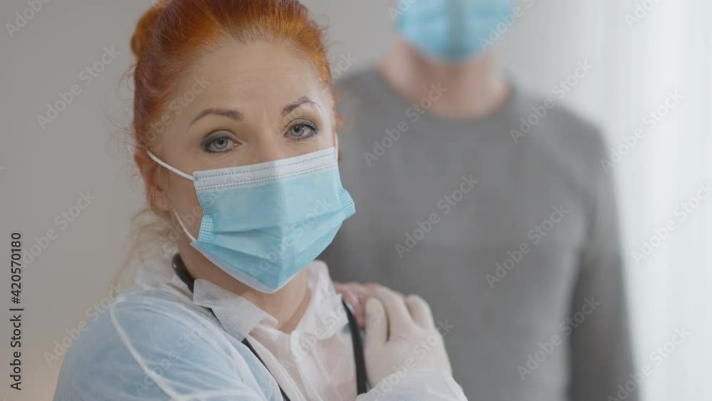 Close-up of frustrated Caucasian woman in doctor uniform and Covid face mask looking at camera as unrecognizable man putting palm on her shoulder. Depressed nurse or physician indoors. Burnout concept Close-up of frustrated Caucasian woman in doctor uniform and Covid face mask looking at camera as unrecognizable man putting palm on her shoulder. Depressed nurse or physician indoors. Burnout concept