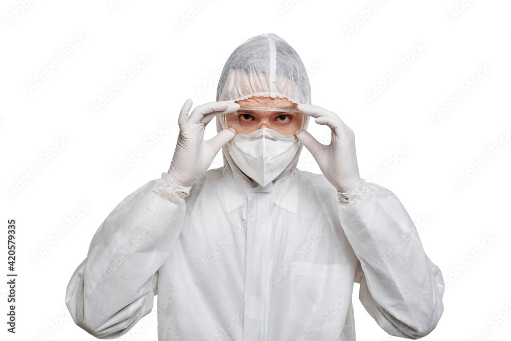A male doctor in PPE puts on protective glasses and looks at the camera. Protection against coronavirus. White isolated background. A male doctor in PPE puts on protective glasses and looks at the camera. Protection against coronavirus. White isolated background.