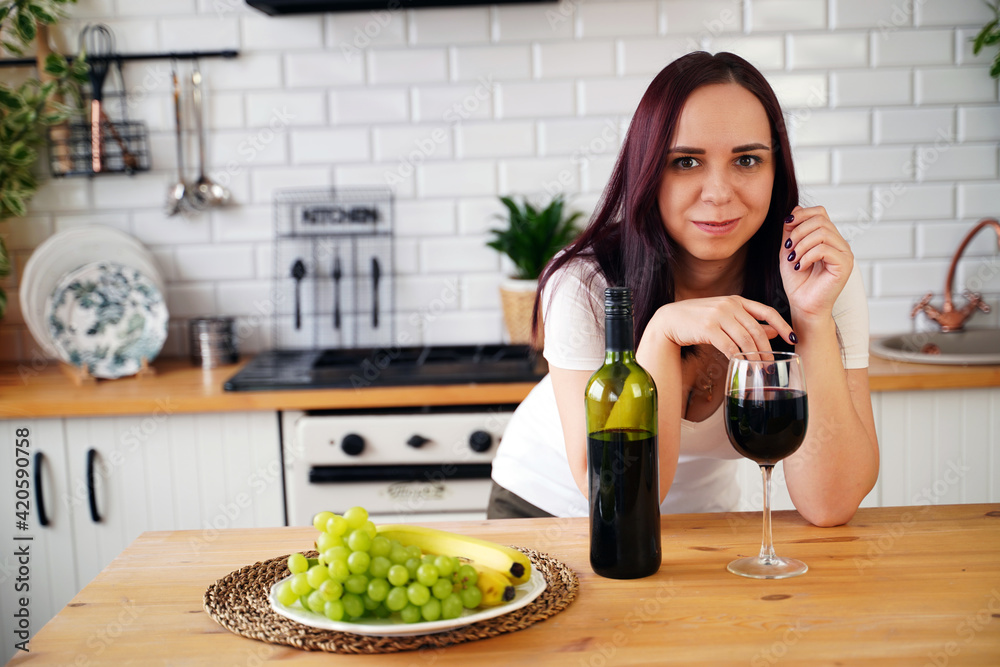 Relaxed young woman drinks red wine, standing at kitchen table. Adult brunette resting with alcohol in kitchen.