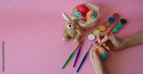 Child`s hands paint Easter egg next to the fluffy toy rabbit, the nest with handmade colored eggs, multicolored paints and art brushes on a pink background. 