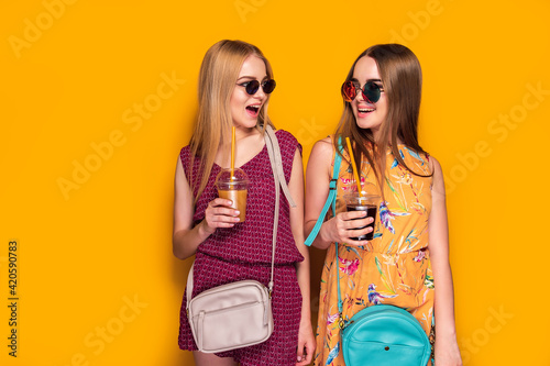 Two cheerful young women girls friends in summer clothes with bags, holding beverages isolated on yellow background.