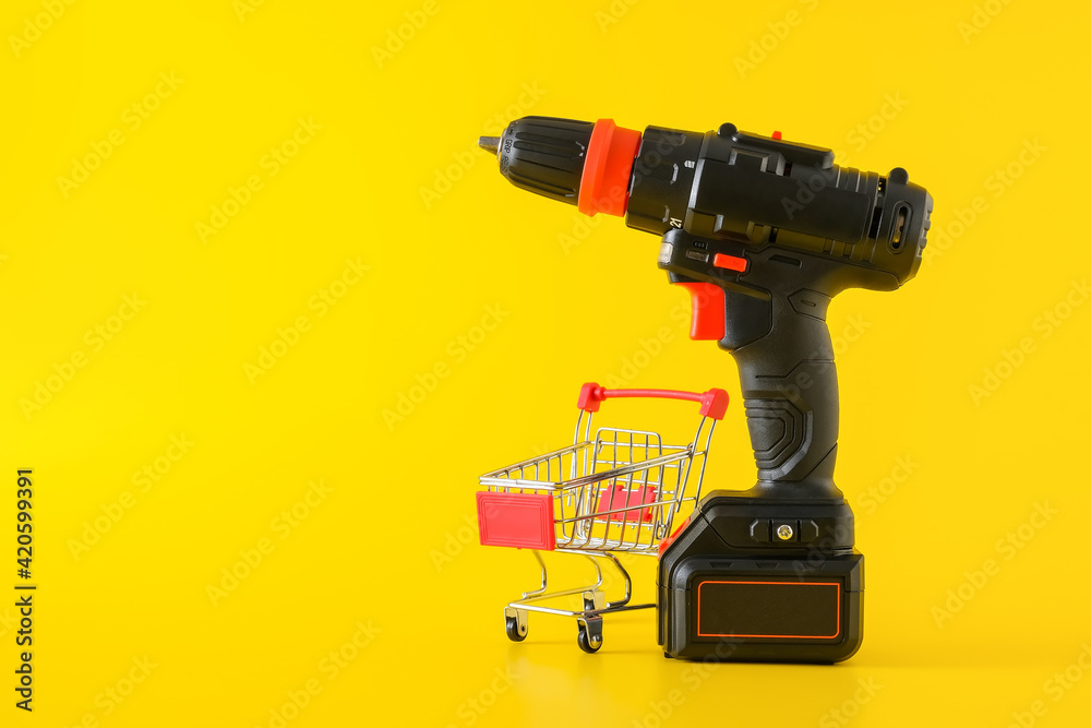 Modern black cordless screwdriver, drill with empty shopping cart on yellow background, copy space for text Modern black cordless screwdriver, drill with empty shopping cart on yellow background, copy space for text