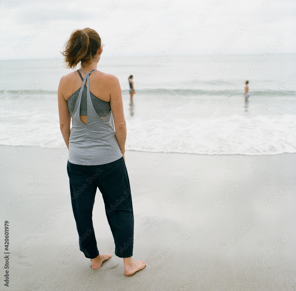 Mother standing on the beach looking at her kids playing in the ocean Mother standing on the beach looking at her kids playing in the ocean