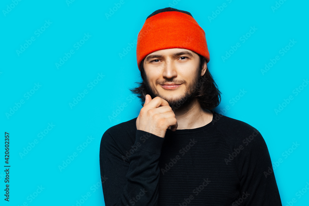Studio portrait of confident young man wearing orange hat and black sweater on blue background.