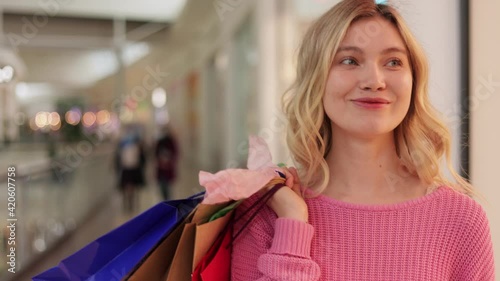 Blonde female in pink sweater walks around shopping mall with colorful shopping bags. Smiling girl looking to the side with colorful bags in hands after successful shopping at store.