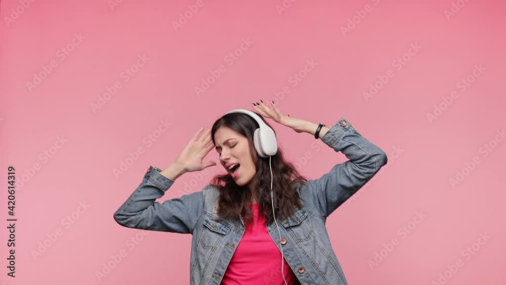 Brunette Female in casual denim jacket pink t-shirt isolated on pink background in studio.People lifestyle concept. Girl listen music in headphones dancing fooling around have fun gesticulating hands. Brunette Female in casual denim jacket pink t-shirt isolated on pink background in studio.People lifestyle concept. Girl listen music in headphones dancing fooling around have fun gesticulating hands.