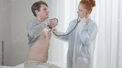 Concentrated woman listening internal sounds as man sitting on medical couch in hospital ward. Caucasian doctor using stethoscope for chest auscultation as patient inhaling and exhaling. photo