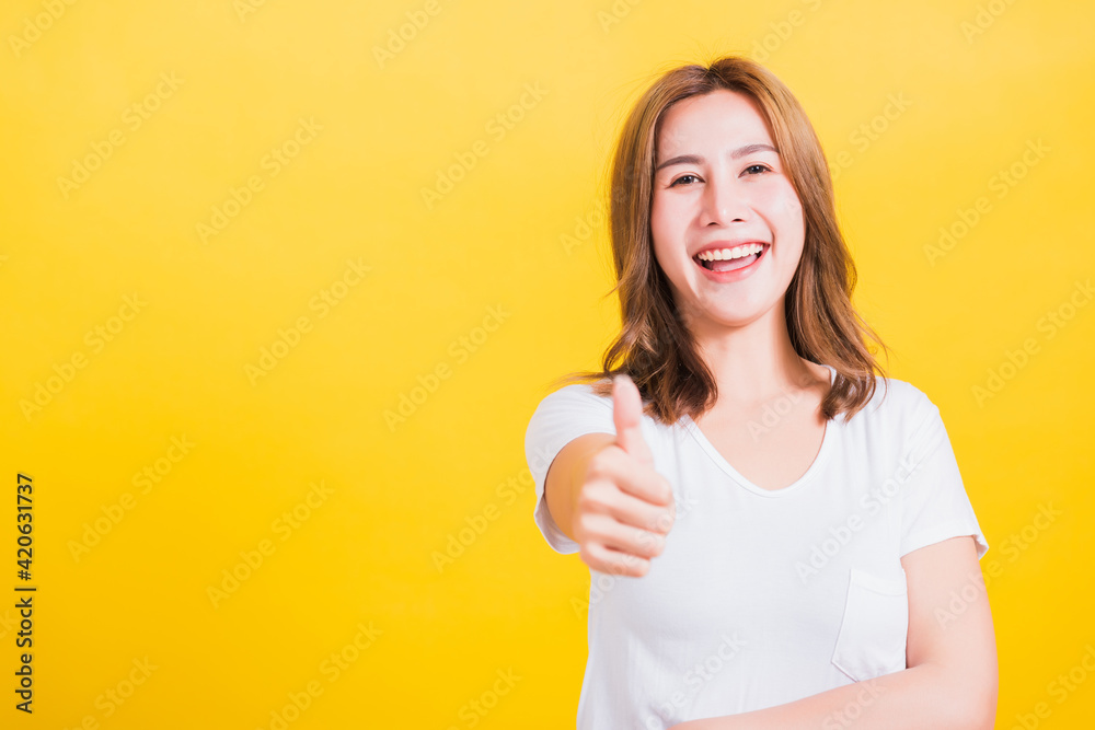 Portrait Asian Thai beautiful happy young woman smiling wear white t-shirt standing successful woman giving one thumbs up gesture sign in studio shot, isolated on yellow background with copy space Portrait Asian Thai beautiful happy young woman smiling wear white t-shirt standing successful woman giving one thumbs up gesture sign in studio shot, isolated on yellow background with copy space
