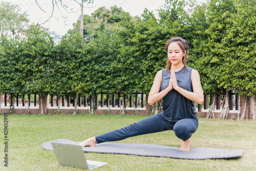 Asian young woman using laptop during online video training practicing yoga outdoors in meditate pose sitting on green grass in nature a field garden park, Stretching health care concept