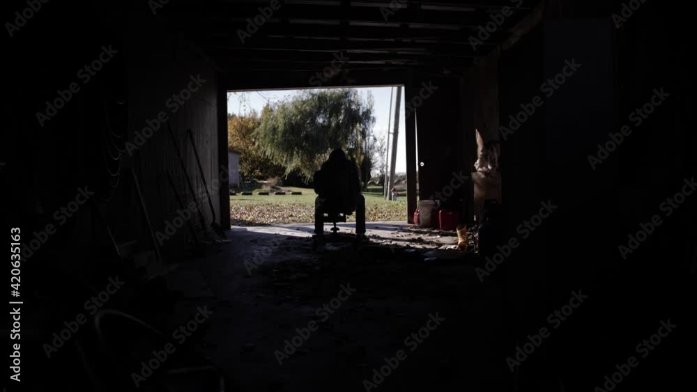 Handheld camera shot of a teenager sitting in a dark and dirty garage wearing a hood. Slow motion 60fps.
