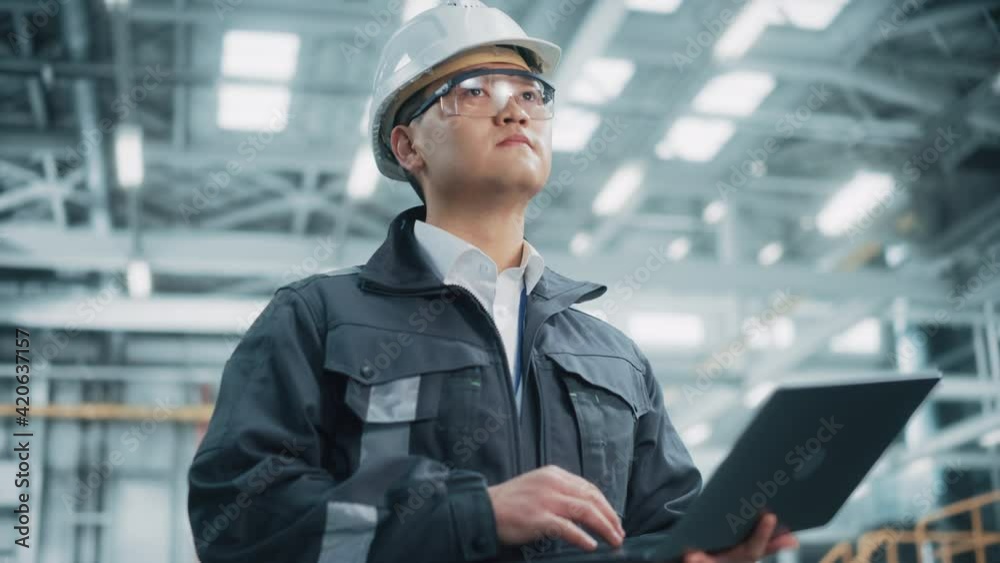 Portrait of a Professional Heavy Industry Asian Engineer/Worker Wearing Safety Uniform and Hard Hat Uses Laptop Computer. Confident Chinese Industrial Specialist Standing in a Factory Facility. Portrait of a Professional Heavy Industry Asian Engineer/Worker Wearing Safety Uniform and Hard Hat Uses Laptop Computer. Confident Chinese Industrial Specialist Standing in a Factory Facility.