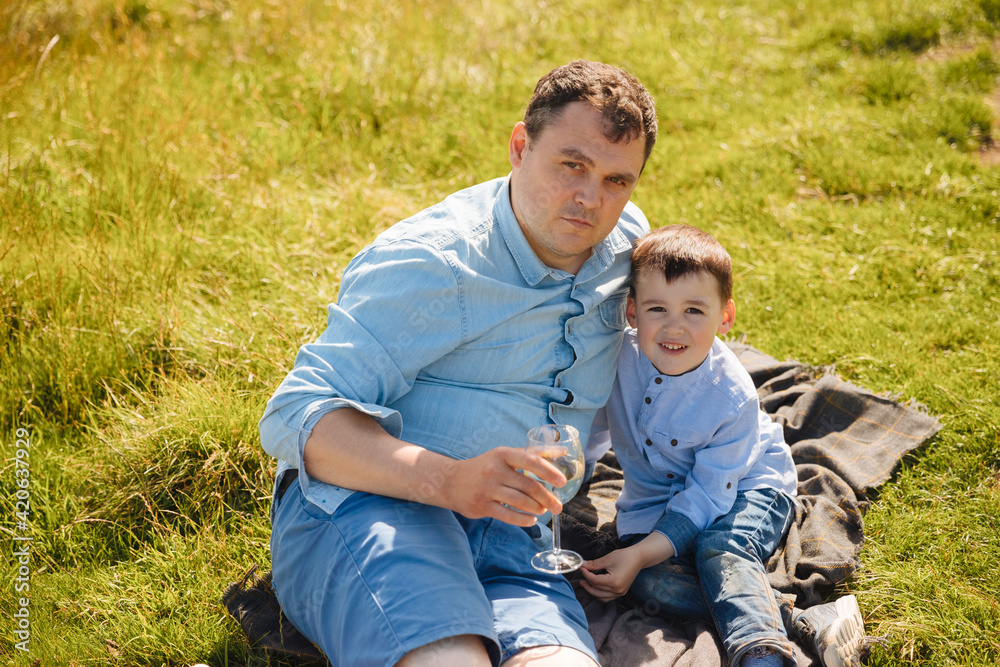 Little boy with his father sitting on a green meadow with the sea in the background. . A child with a parent at the seaside. Family vacation with dad by the sea. Little boy with his father sitting on a green meadow with the sea in the background. . A child with a parent at the seaside. Family vacation with dad by the sea.