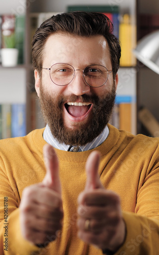 Man thumb up. Positive laughing bearded man in glasses in office or apartment room looking at camera and shows his two hands thumb up. Close-up