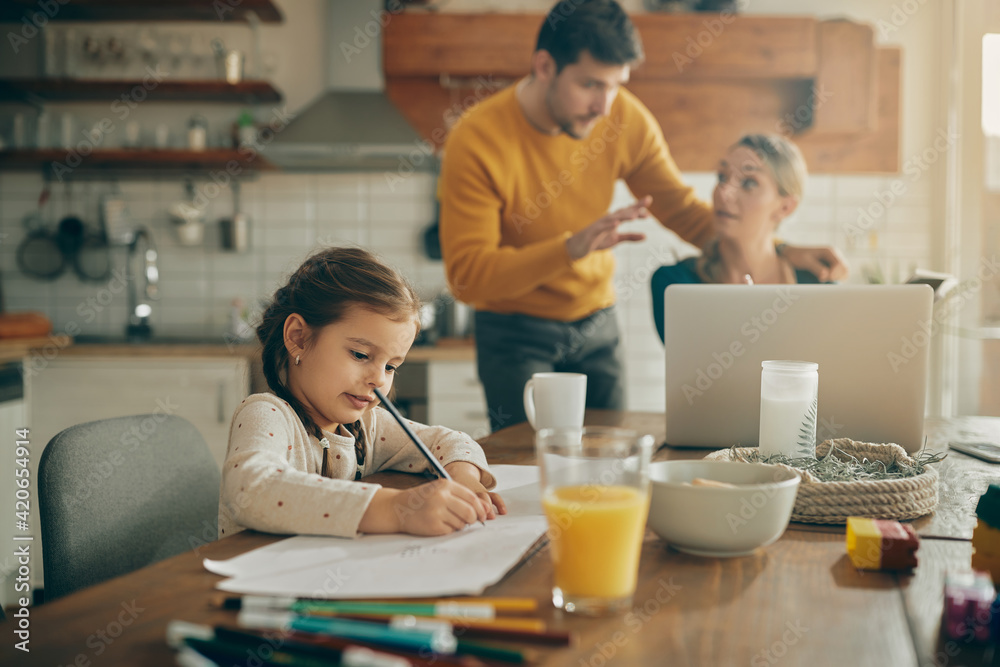Small girl writing homework while parents are working in the background at home. Small girl writing homework while parents are working in the background at home.