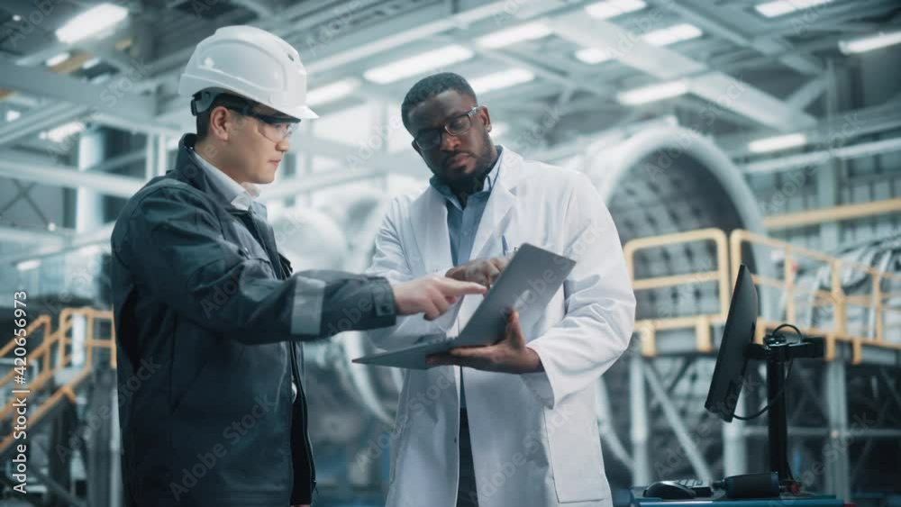 Team of Diverse Professional Heavy Industry Engineers Wearing Safety Uniform and Hard Hat Working on Laptop Computer. African American Technician and Asian Worker Talking on a Meeting in a Factory. Team of Diverse Professional Heavy Industry Engineers Wearing Safety Uniform and Hard Hat Working on Laptop Computer. African American Technician and Asian Worker Talking on a Meeting in a Factory.