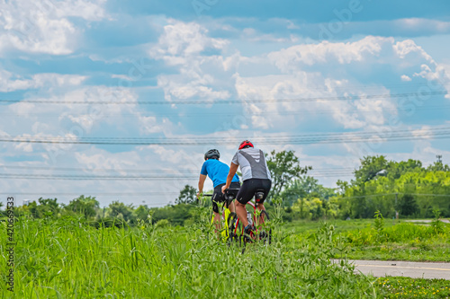 Two high energy colorfully dressed cyclists prepare to pass under high energy wires while on a nature cycle path. The cycle path is framed by green fields and a blue cloud filled sky
