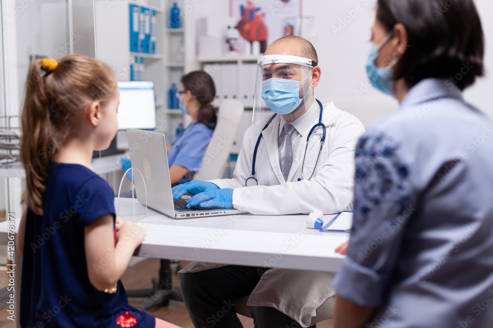 Obraz premium Caring doctor discussing symptoms with mother in medical clinic. Specialist in pediatry medicine with protection mask and visor against covid19 talking about diagnostic and healtcare treatment