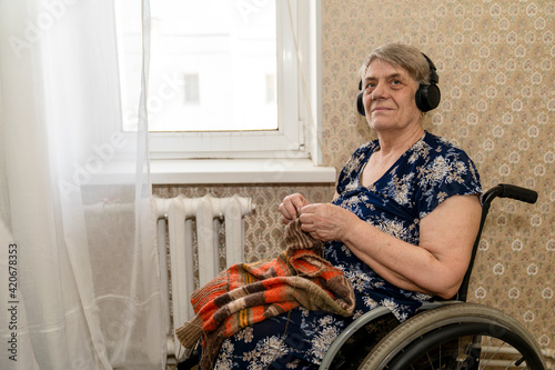 an elderly woman with headphones sits in a wheelchair and, with a slight smile on her face, knits a woolen thing on the needles