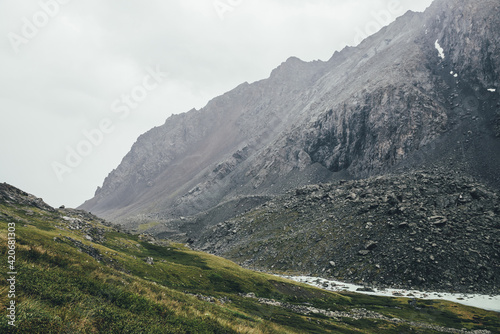 Atmospheric alpine landscape with narrow valley with mountain creek and sharp rocks under gray sky. Bleak highlands scenery with pointed rockies on mountainside and mountain creek in overcast weather.