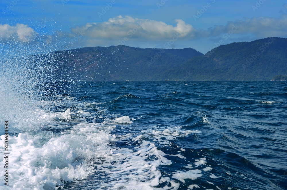 Seawater splash from speed boat, suspended in the air with blue sky and mountain in background.