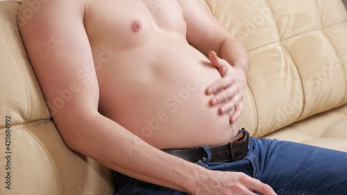 Unrecognizable man in jeans and no t-shirt stroking swollen belly while sitting on couch.