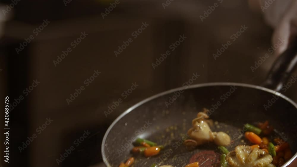 A close video of the frying octopuses with vegetables on a hot pan which is throwing up a cook by hand in a disposable glove in the kitchen. Steam puffs over the dish.