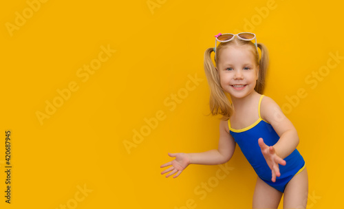 Beautiful cute girl with ponytails and sunglasses on her head on a yellow background. A funny blonde little girl in a blue bathing suit bent merrily to the side. Horizontal banner with place for text.