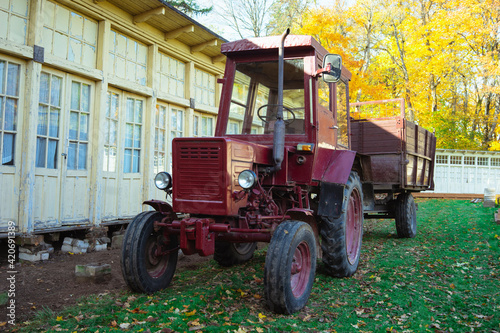 Vintage red tractor parked on green grass near the house in autumn. Trailer for transporting heavy loads on the terrain. Latvia