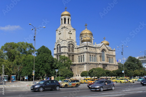 On the opposite side of the road is an ancient beige temple with golden domes against the blue sky