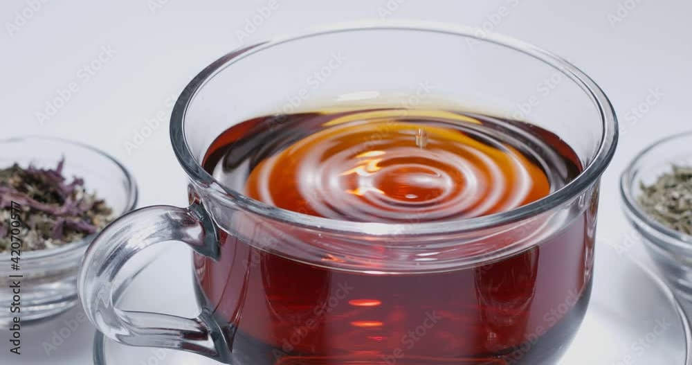 Drop slowly falls into a glass cup of tea. Around there are small bowls with different types of leaf tea. White background. Drop slowly falls into a glass cup of tea. Around there are small bowls with different types of leaf tea. White background.