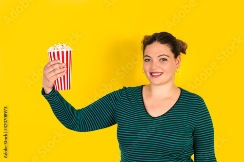 Mujer joven sonriente y alegre sosteniendo un cubo de palomitas de maíz con el brazo levantado sobre un fondo amarillo liso y aislado. Visat de frente. Copy space photo