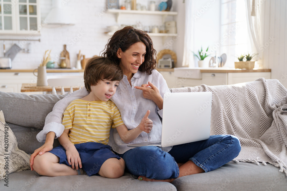 Family, lockdown concept. Happy mother and kid son greeting online looking at web camera using laptop for video call, sitting on couch at home. Smiling mom and child having fun talking in video chat.