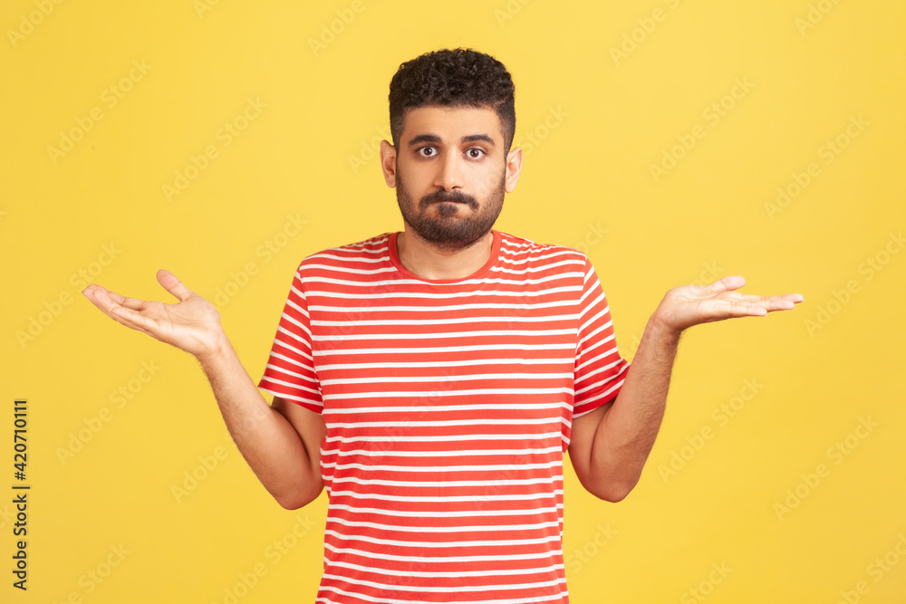 Puzzled clueless man with beard in striped red t-shirt spreading hands and shrugging shoulders, not sure and indifferent in some questions. Indoor studio shot isolated on yellow background Puzzled clueless man with beard in striped red t-shirt spreading hands and shrugging shoulders, not sure and indifferent in some questions. Indoor studio shot isolated on yellow background