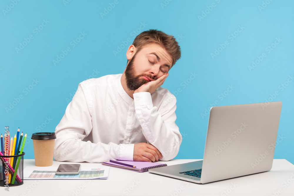 Bored sleepy man office worker napping sitting at workplace with laptop on desk leaning head on hand, feeling boredom, lazy to work, overworking. Indoor studio shot isolated on blue background Bored sleepy man office worker napping sitting at workplace with laptop on desk leaning head on hand, feeling boredom, lazy to work, overworking. Indoor studio shot isolated on blue background