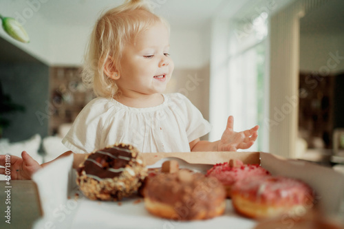 cute beautiful little girl with blonde blue eyes eats delicious fresh donuts. Happy childhood  looks away. She s wearing a comfortable white T-shirt.
