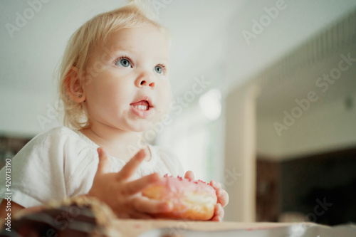 cute beautiful little girl blonde blue eyes eating a donut. Happy childhood, looks away. Close portrait, copy space. She's wearing a comfortable white T-shirt.