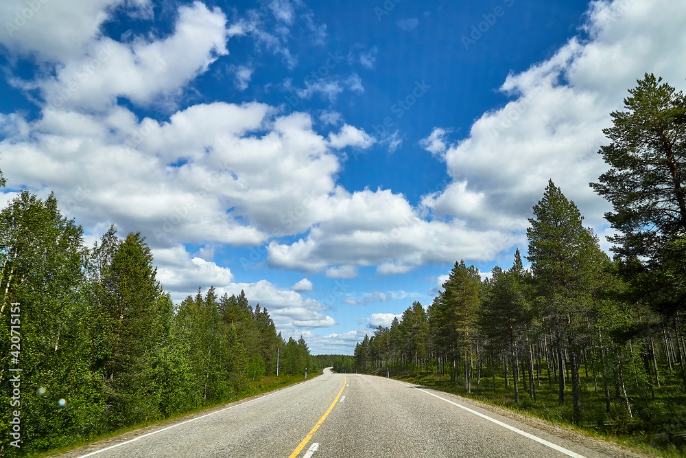 Fototapeta premium View from relief car windscreen on the blue sky with white clouds, grey asphalt road and landscape with forest and green teeses. Landscape through window