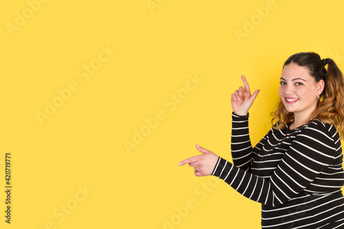 Canvastavla Mujer emocionada y sonriente mirando a la cámara mientras apunta con los dedos hacia un costado sobre un fondo amarillo liso y aislado