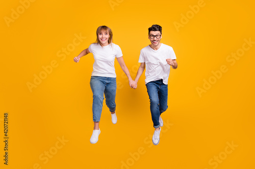 Full length photo of funny sweet two persons dressed white t-shirt running jumping holding arms isolated yellow color background