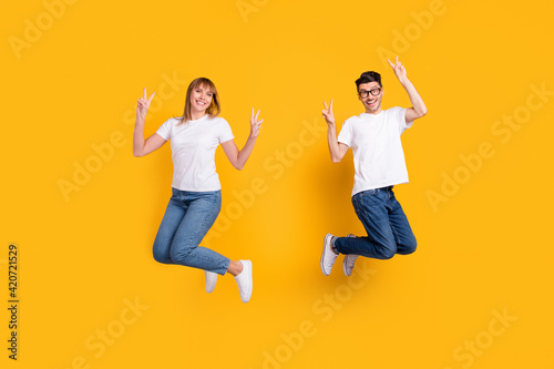 Full length photo of sweet pretty two persons dressed white t-shirt jumping high showing v-signs isolated yellow color background