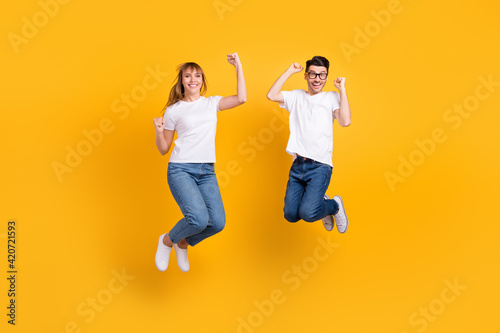 Full length photo of cute lucky couple wear white t-shirt spectacles holding hands jumping rising fists isolated yellow color background