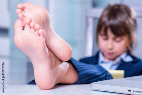 Humorous portrait of cute little business child girl with bare feet works with laptop. Selective focus on bare feet. photo