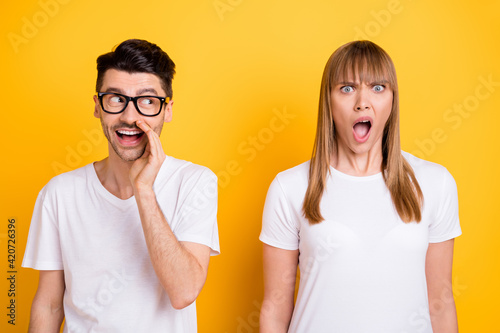 Photo of shocked funny young couple wear white t-shirt spectacles arm lips telling secret isolated yellow color background