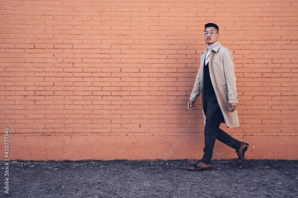 Middle-aged Latino man with glasses, beige trench coat, and brick background poses Middle-aged Latino man with glasses, beige trench coat, and brick background poses